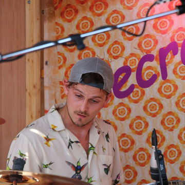 Schlagzeuger mit Basecap und Blumenhemd vor dem secret stage Schriftzug, Kieler Woche 2023